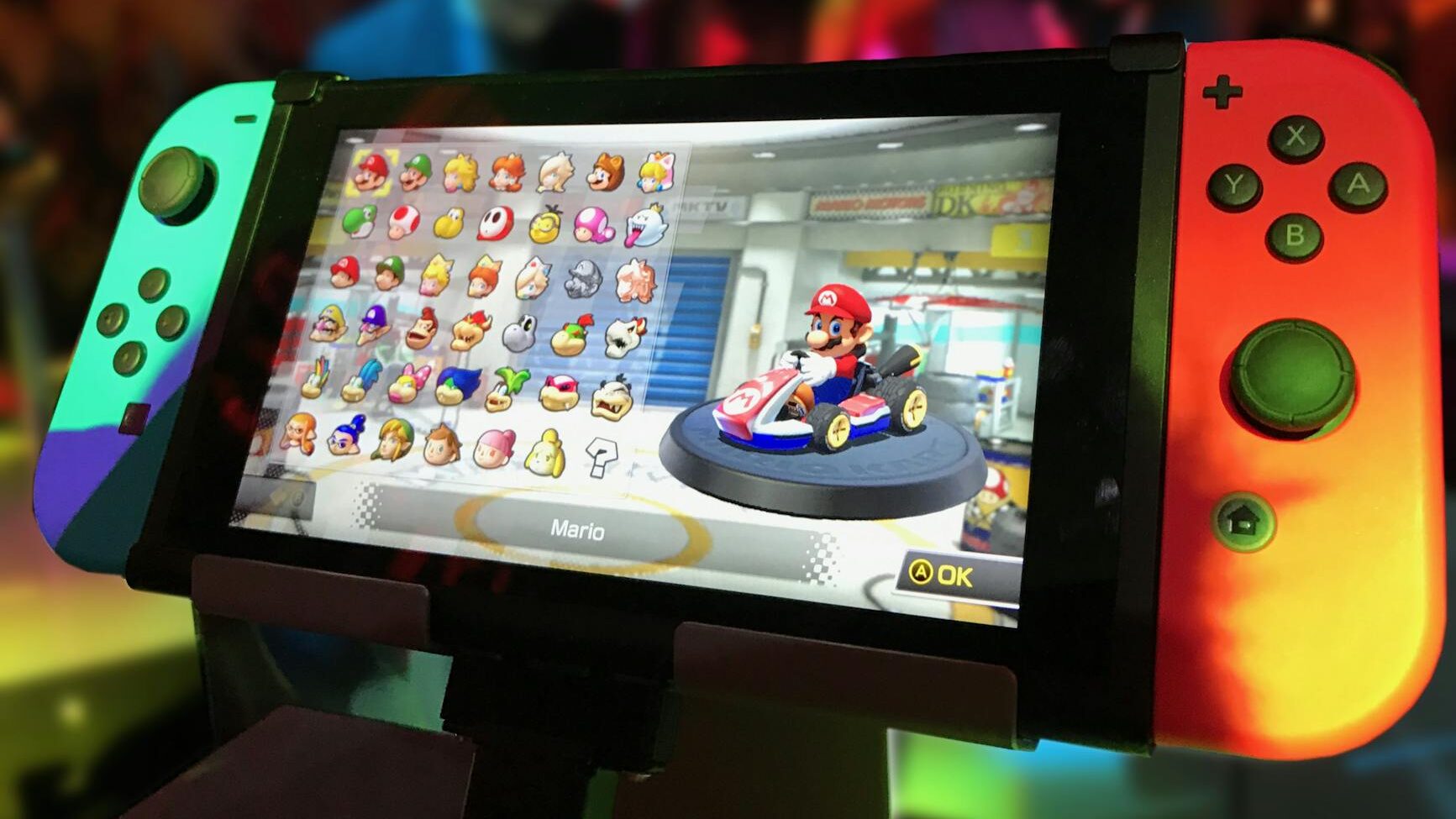 Close-up of a Nintendo Switch showing Mario Kart selection screen, gaming atmosphere in the background.