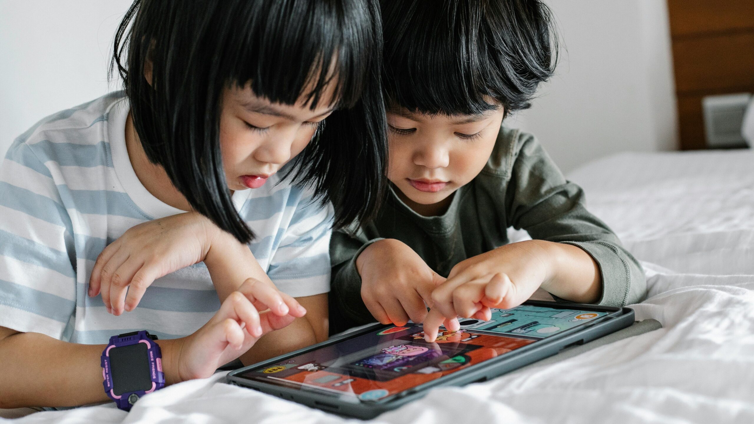 Attentive Asian boy and girl playing video game on modern tablet while lying on bed in bedroom in morning time